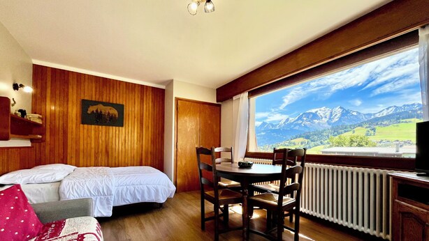 A room with a bed, a dining table with chairs, a radiator, and a large window with a mountain view.