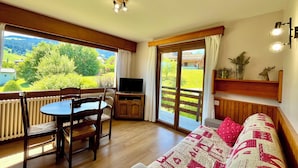 A room with a wooden floor, a sofa with red and white patterned cushions, a dining table with chairs, a television on a wooden stand, and a large sliding glass door leading to a balcony with a view of greenery and a building.