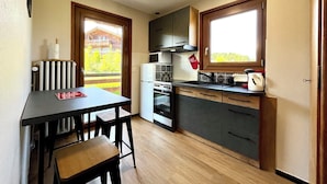 A compact kitchen with a dining area, wooden cabinets, a refrigerator, and a window providing a view of a wooden building outside.