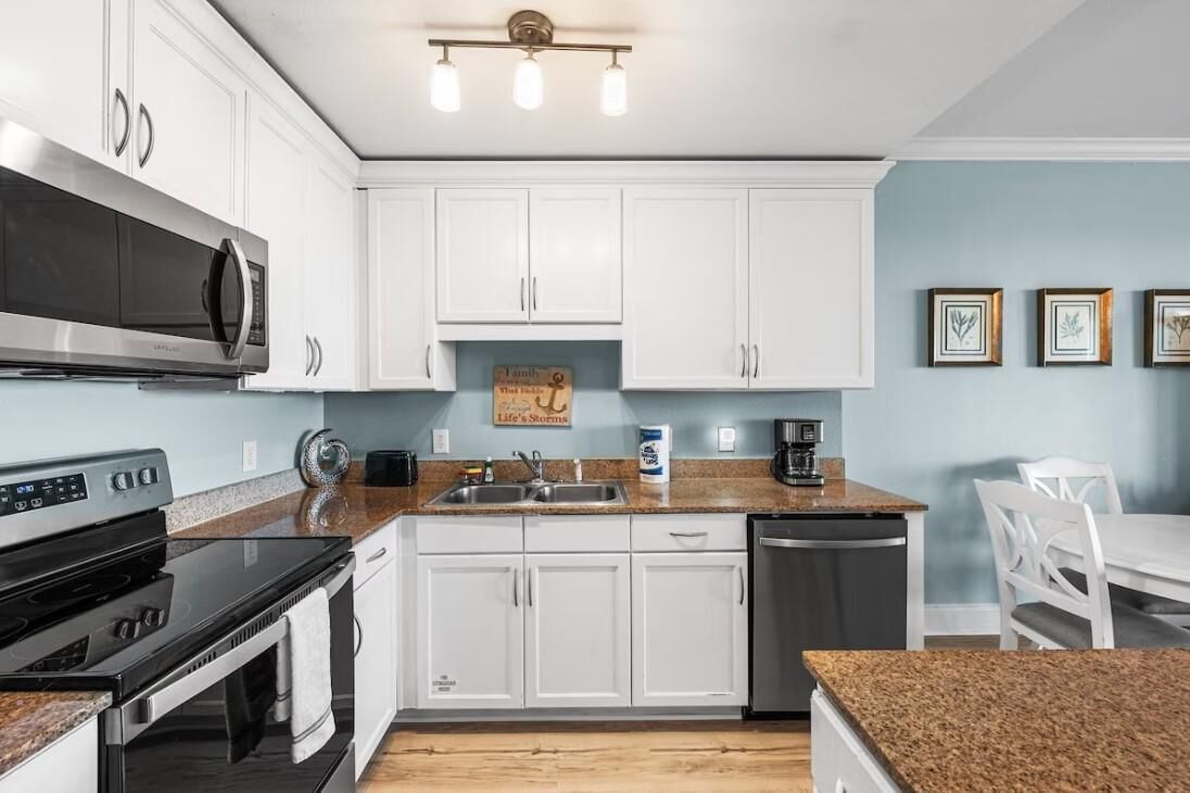 A modern kitchen with white cabinets, a stainless steel microwave, a stove, and a dishwasher. There is a sink with a faucet, a coffee maker, and a framed picture on the wall.