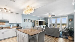 A modern kitchen with white cabinets, a granite countertop, and a chandelier. There is a dining area with a gray sofa and a large window offering a view of the outdoors. - Laketown Wharf 1933 (Panama City Beach)