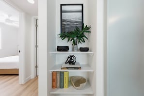 A white room with a bed, a shelf with books, a plant, and a framed picture.