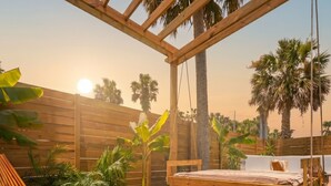 Una terraza de madera con un columpio, un banco y una cama con vistas a palmeras.