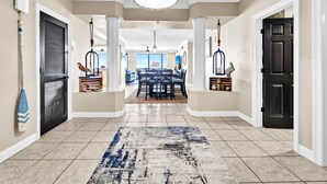 A spacious hallway with a dining area in the background, featuring a chandelier, decorative birdcages, and a patterned rug.