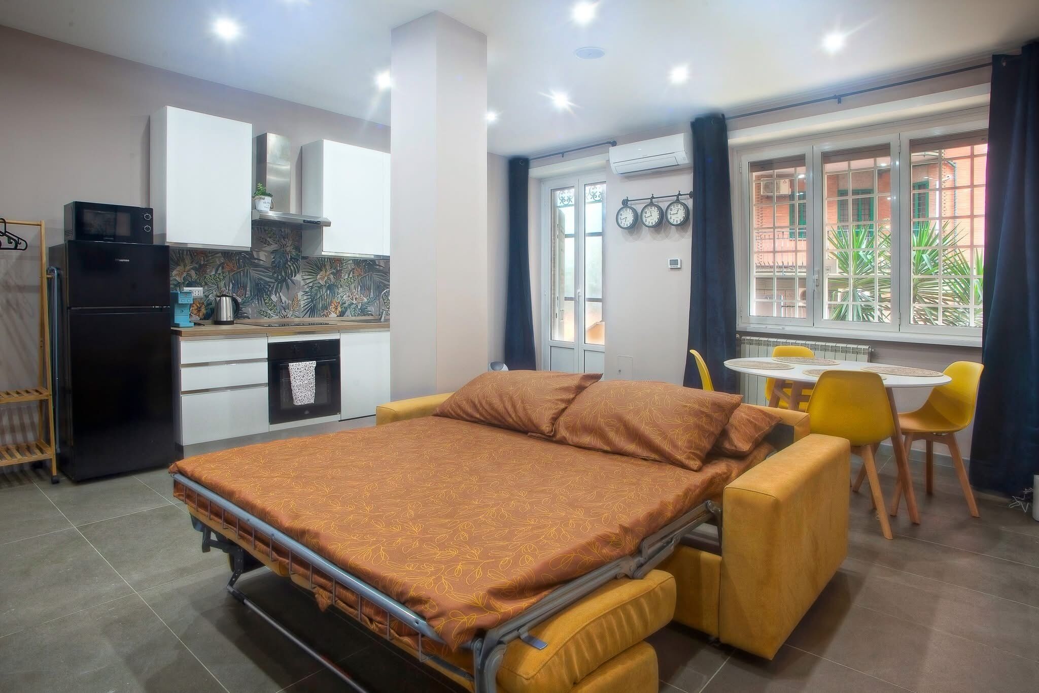 Een kleine kamer met een bed, kitchenette, eethoek en een raam met gordijnen.