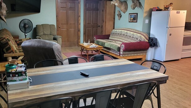 A living room with a wooden dining table, chairs, a sofa, a television, and a refrigerator.