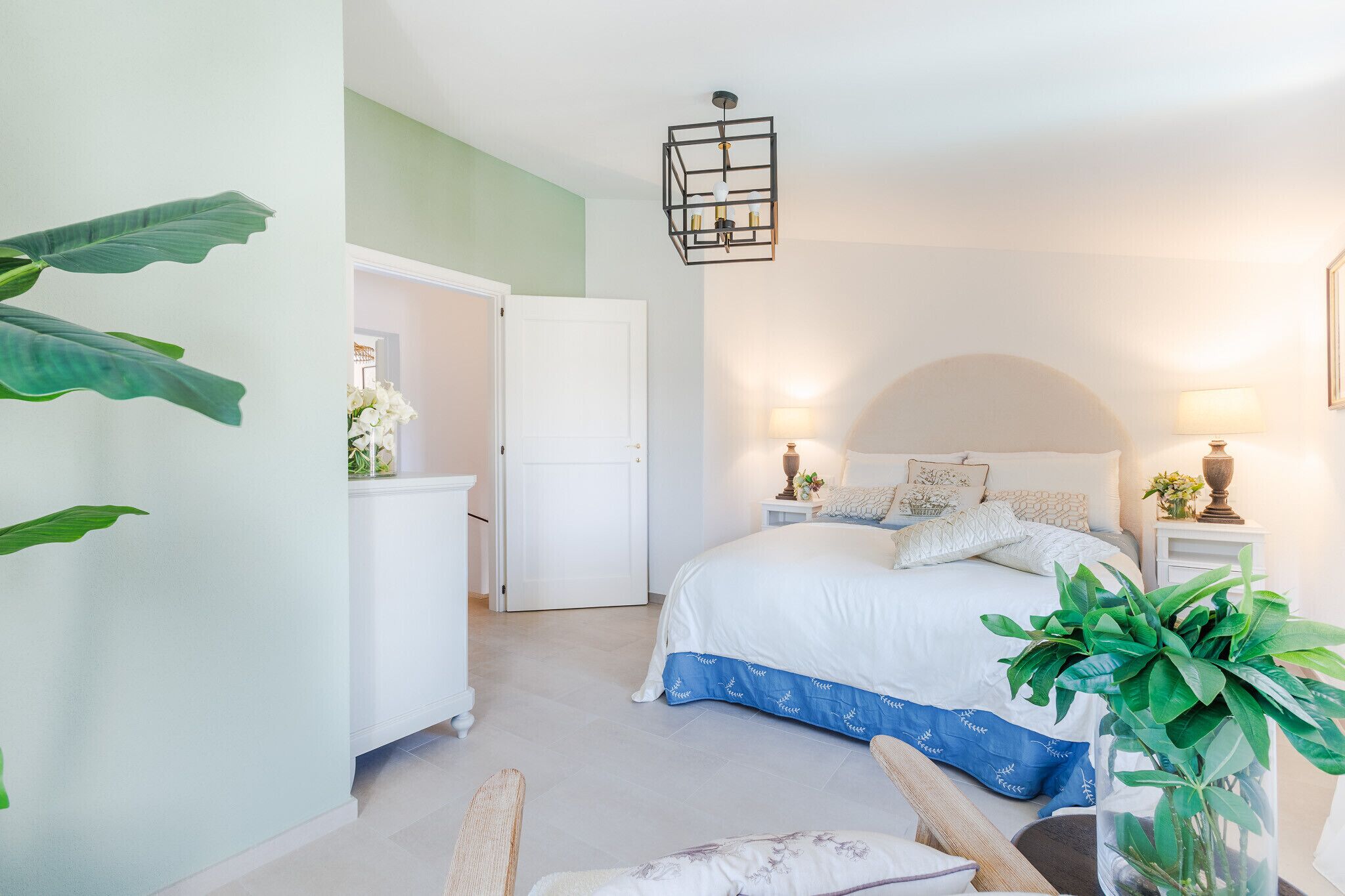 Quarto com cama, mesas de cabeceira, luminária pendente, cadeira e planta em vaso.
