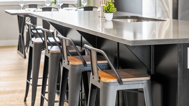 A modern kitchen with a long island counter, bar stools, and a sink. - Cowboy Chalet SE, AC, Games, King Bed, 20 min to Down Town (Calgary)