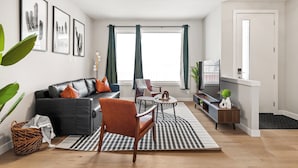 A modern living room with a sofa, armchair, coffee table, and a TV. There are framed pictures on the wall, a plant, and a basket on the floor. - Cowboy Chalet SE, AC, Games, King Bed, 20 min to Down Town (Calgary)