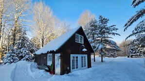 Une cabane en bois dont le toit est recouvert de neige, entourée d’arbres chargés de neige et sous un ciel dégagé.