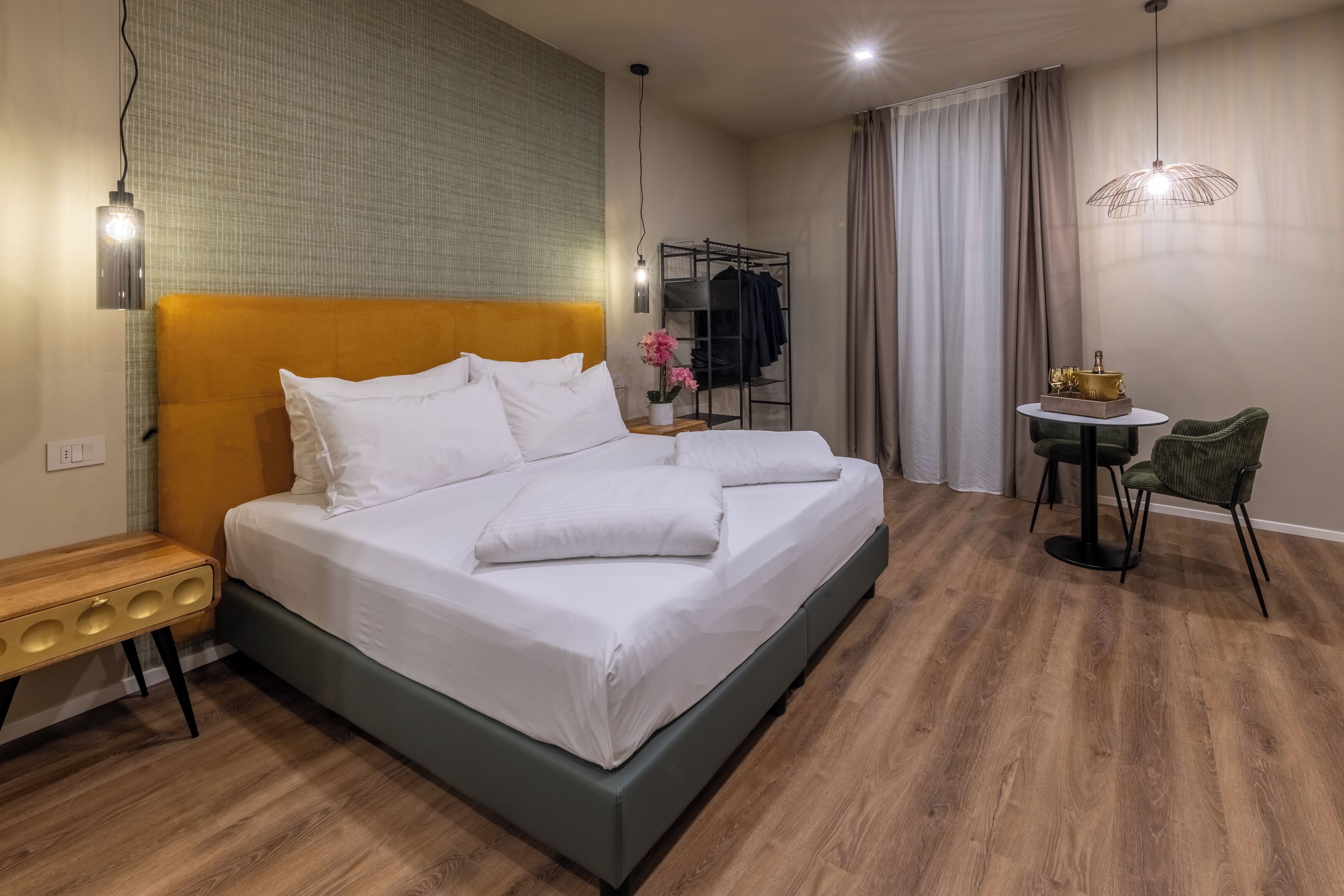 A modern hotel room with a large bed, a small table with chairs, a hanging light fixture, and a coat rack.