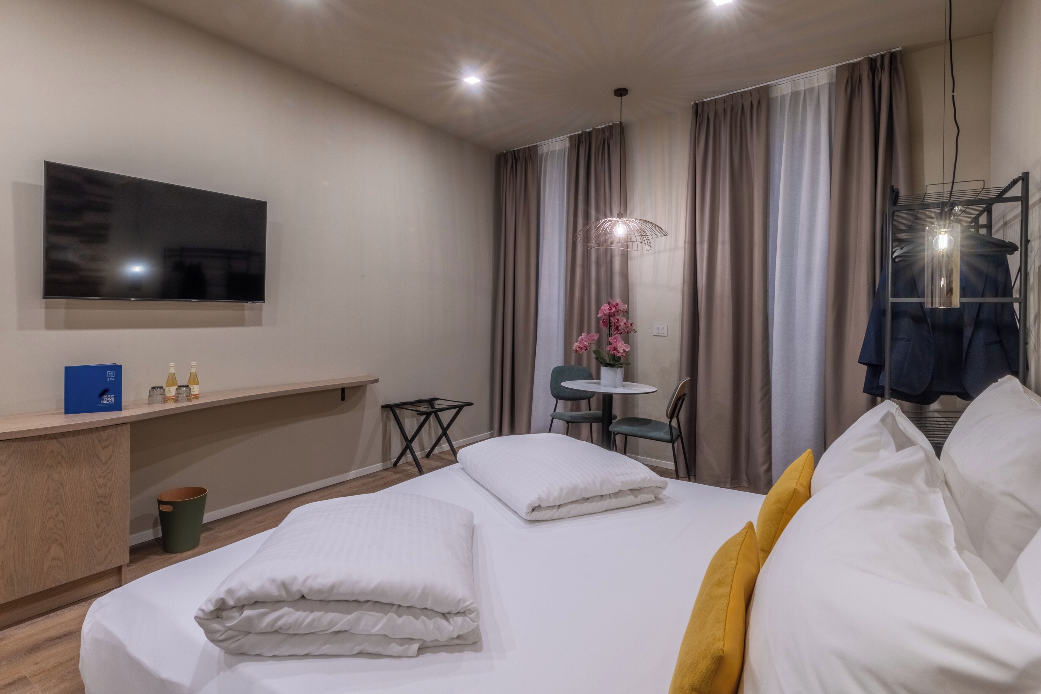 Habitación de hotel con cama, televisión montada en la pared, una pequeña mesa con sillas y una lámpara colgante.