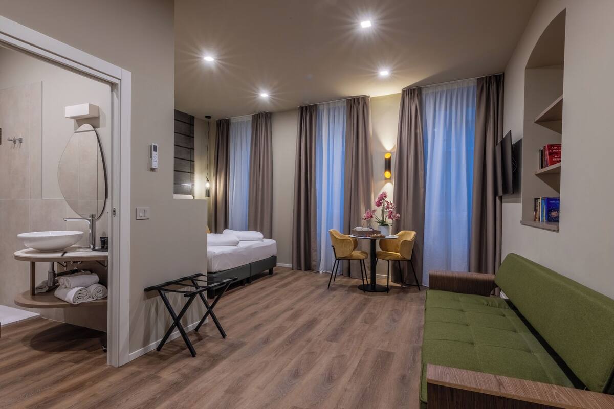 A modern hotel room with a wooden floor, a bed, a green sofa, a round table with chairs, a small round table with a vase, and a TV on the wall.