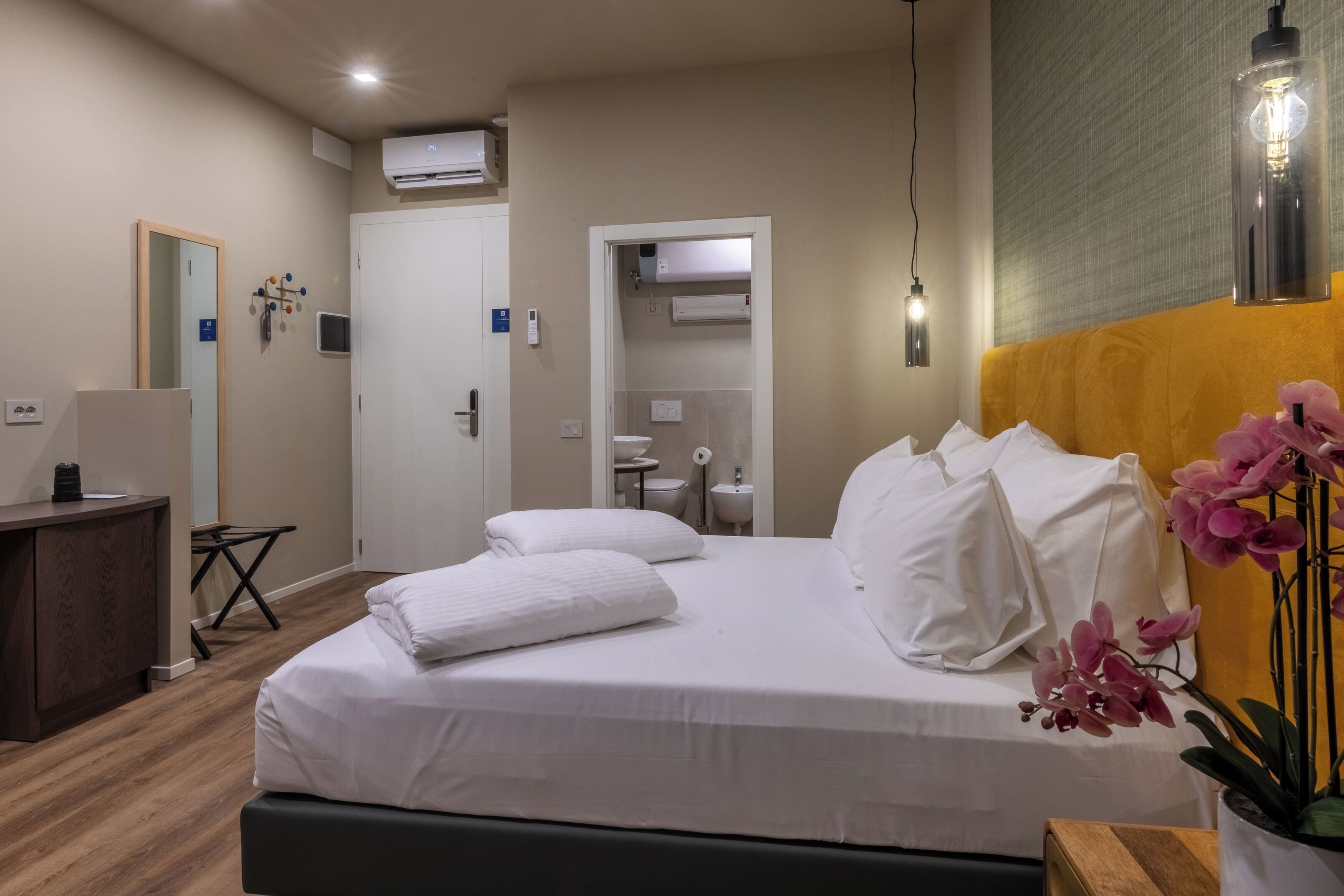 A hotel room with a bed, bedside table, and a mirror. There is a bathroom visible through an open door.