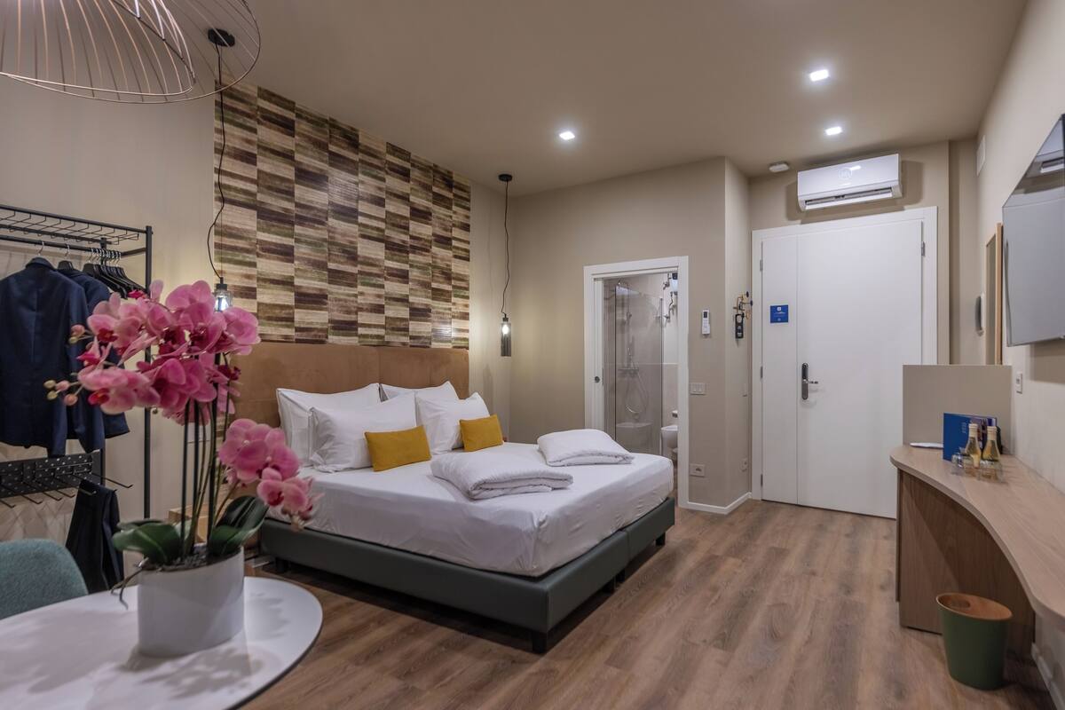 A modern hotel room with a bed, a bedside table with a potted orchid, a shower area, and a small desk with a mirror and toiletries.
