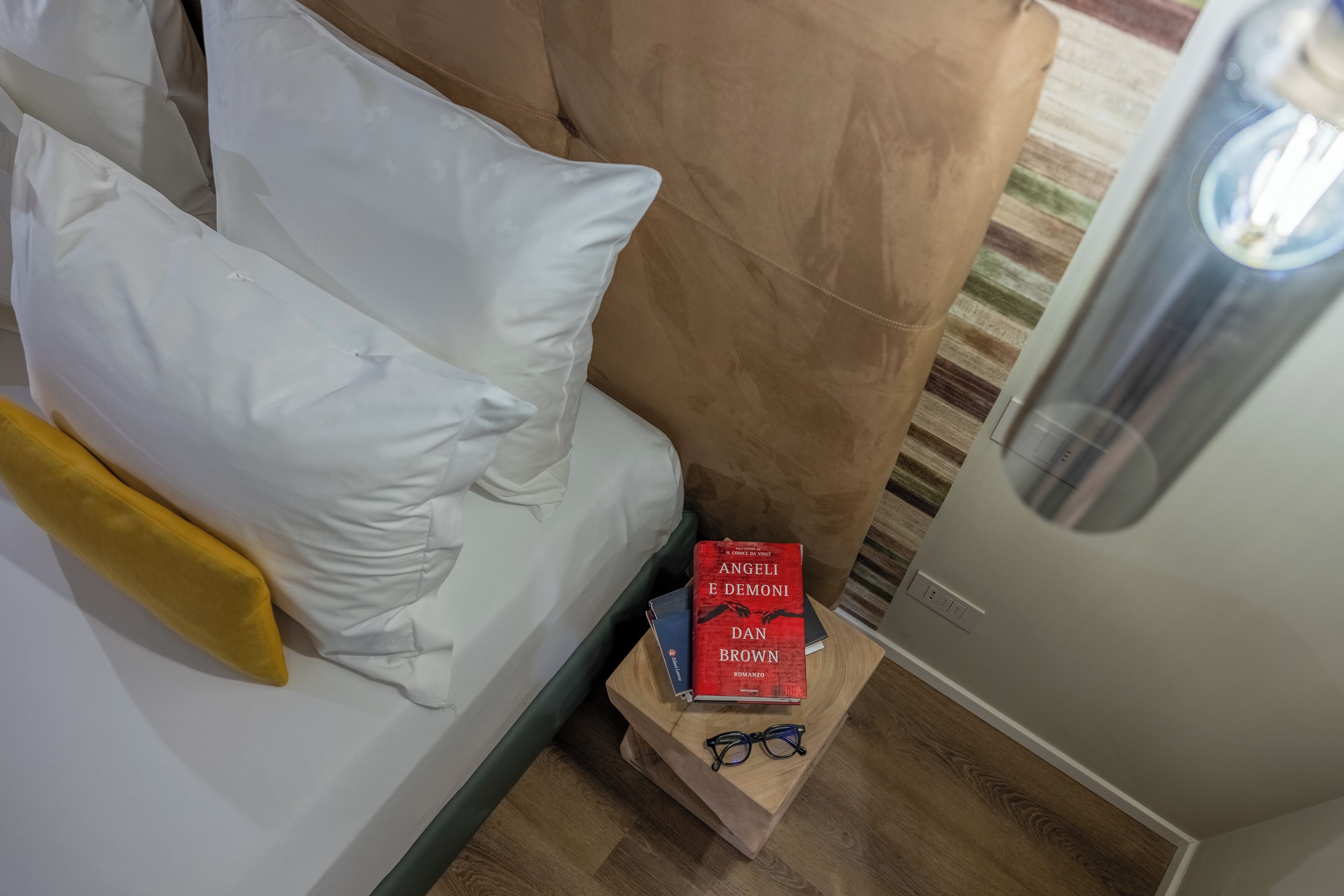 Ein Schlafzimmer mit einem Bett, Kissen, einem Nachttisch mit Büchern und Brillen sowie einer Lampe.