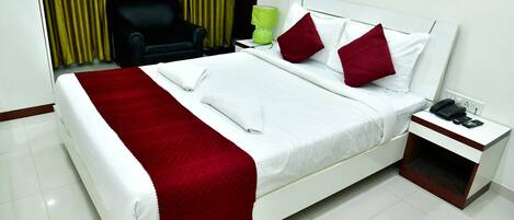 A neatly made bed with white linens and red accents, a bedside table with a phone and charger, and a black chair in the background.