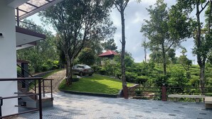 A paved courtyard with a car parked in the background, surrounded by lush greenery and trees.