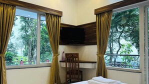 A hotel room with a bed, a desk with a flat-screen TV, a chair, and large windows with curtains.