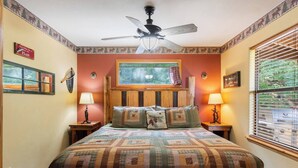 A bedroom with a large bed, a ceiling fan, two bedside lamps, a window with blinds, and a wall with a deer decoration. - River View, Sleeps 6, Hot Tub (Carroll County)