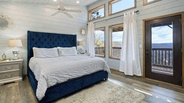 A bedroom with a large bed, a ceiling fan, a nightstand, and a view of the outdoors through a door.