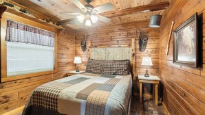 Acogedora habitación de cabaña de madera con cama, dos mesitas de noche con lámparas, ventilador de techo y ventana con persianas.