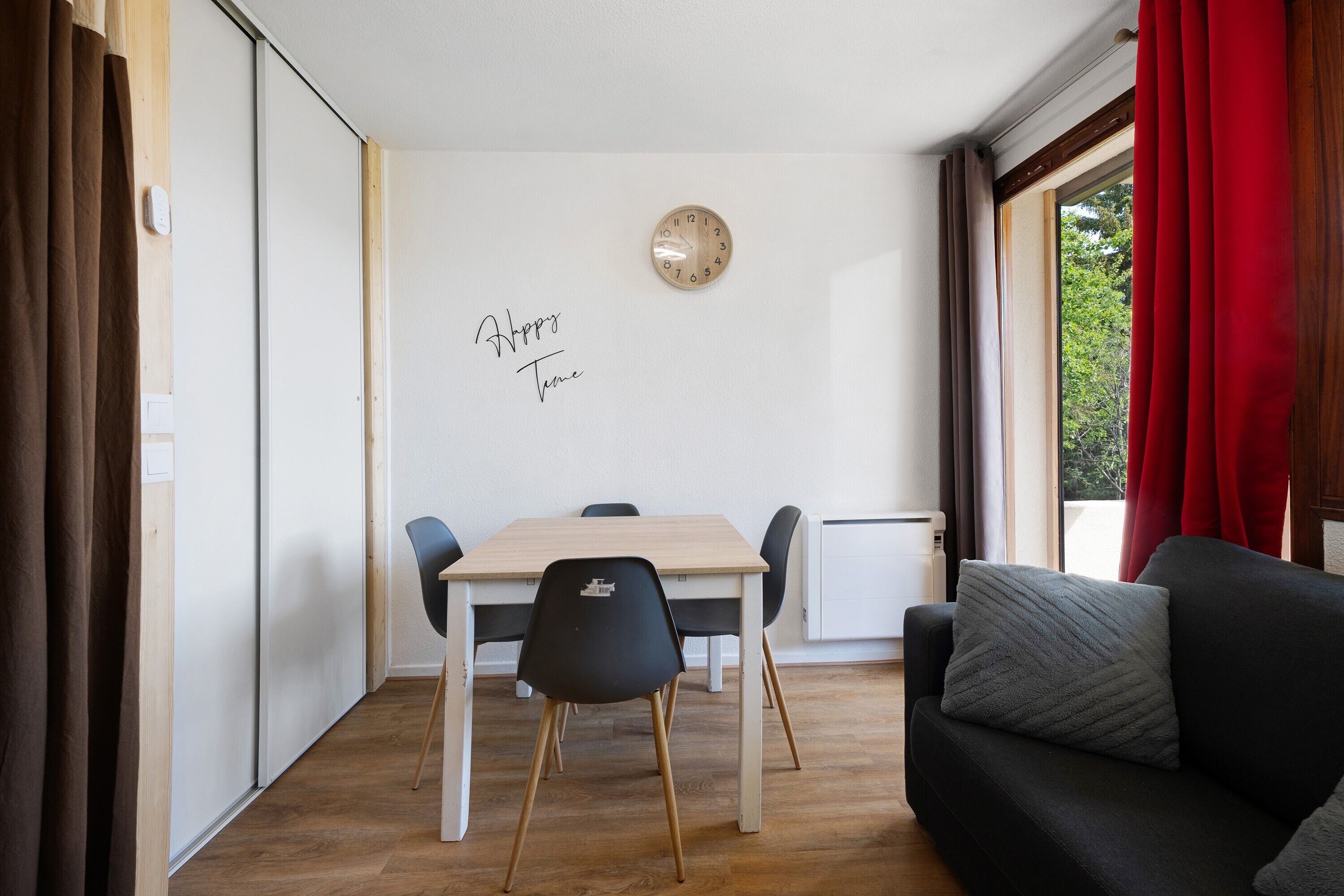 Ein Zimmer mit Esstisch, Stühlen, einer Couch, einer Uhr und einem Fenster mit Vorhängen.