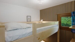 Een stapelbed met wit beddengoed, een nachtkastje met een lamp, een ingelijst beeld aan de muur en een raam met uitzicht op groen.