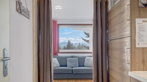 Een kamer met een houten vloer, een grijze bank met kussens, een raam met uitzicht op bergen en een deur met een bordje.