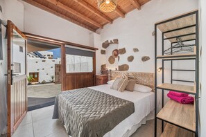 A bedroom with a bed, wooden headboard, stone wall decor, a wooden shelf with a towel, and a view through an open door to an outdoor area.