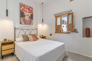 Un dormitorio con cama, mesitas de noche, un cuadro, una ventana y una estantería de pared.