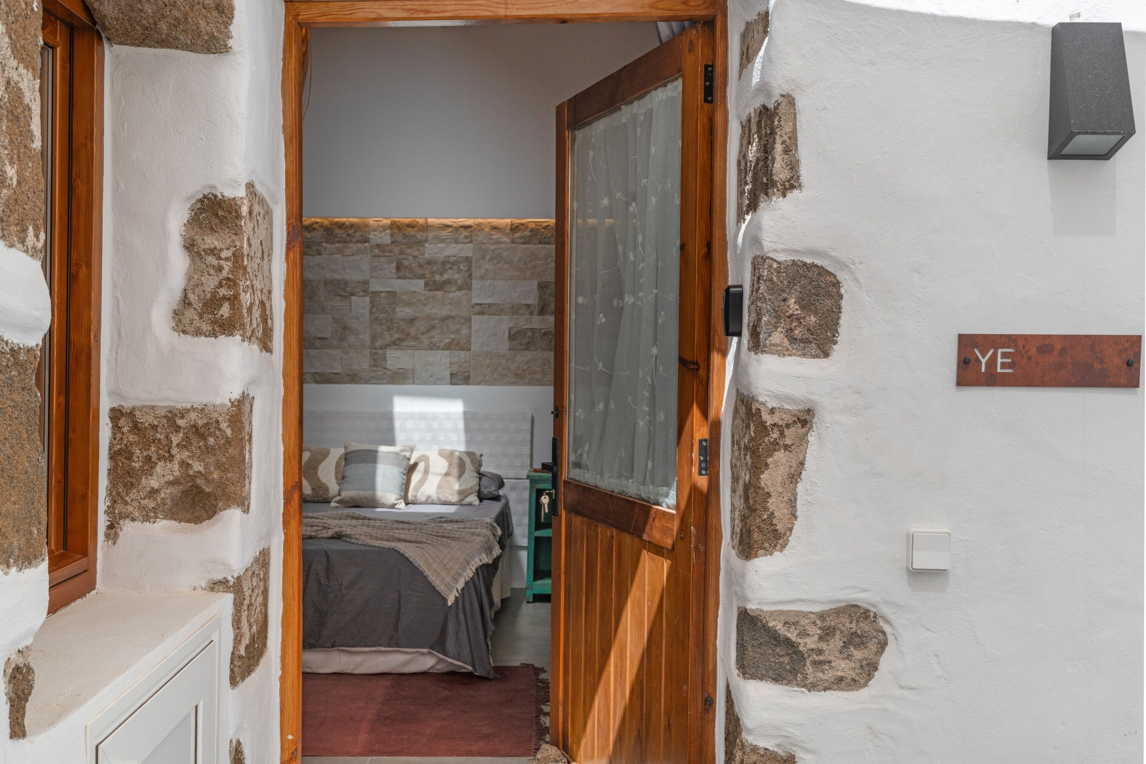 A narrow hallway leading to a bedroom with a stone wall, a wooden door, and a bed with a gray blanket.