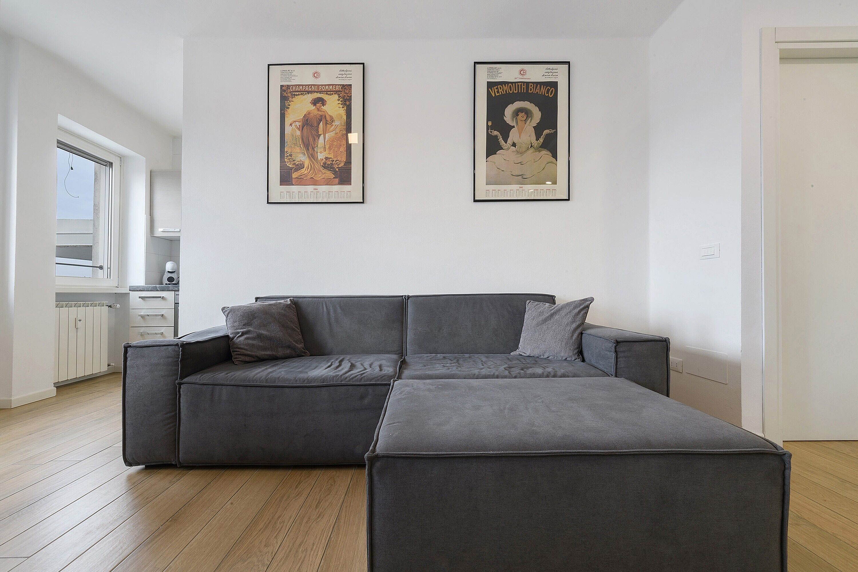Ein modernes Wohnzimmer mit einer Eckcouch, einer Küche im Hintergrund und zwei gerahmten Postern an der Wand.