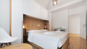 A modern hotel room with a wooden headboard, a bed with white bedding, a white chair, a small table, and a wardrobe. - Room 'Enza 1' with Wi-Fi (Bergamo)