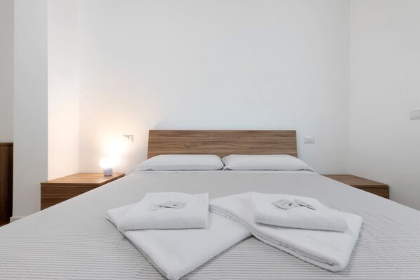 A neatly made bed with white linens and towels, a wooden headboard, and a bedside table with a lamp.