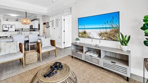 A modern living room with a large flat-screen TV, a dining area with white furniture, and a kitchen in the background. - Tropic Winds 503 (Panama City Beach)