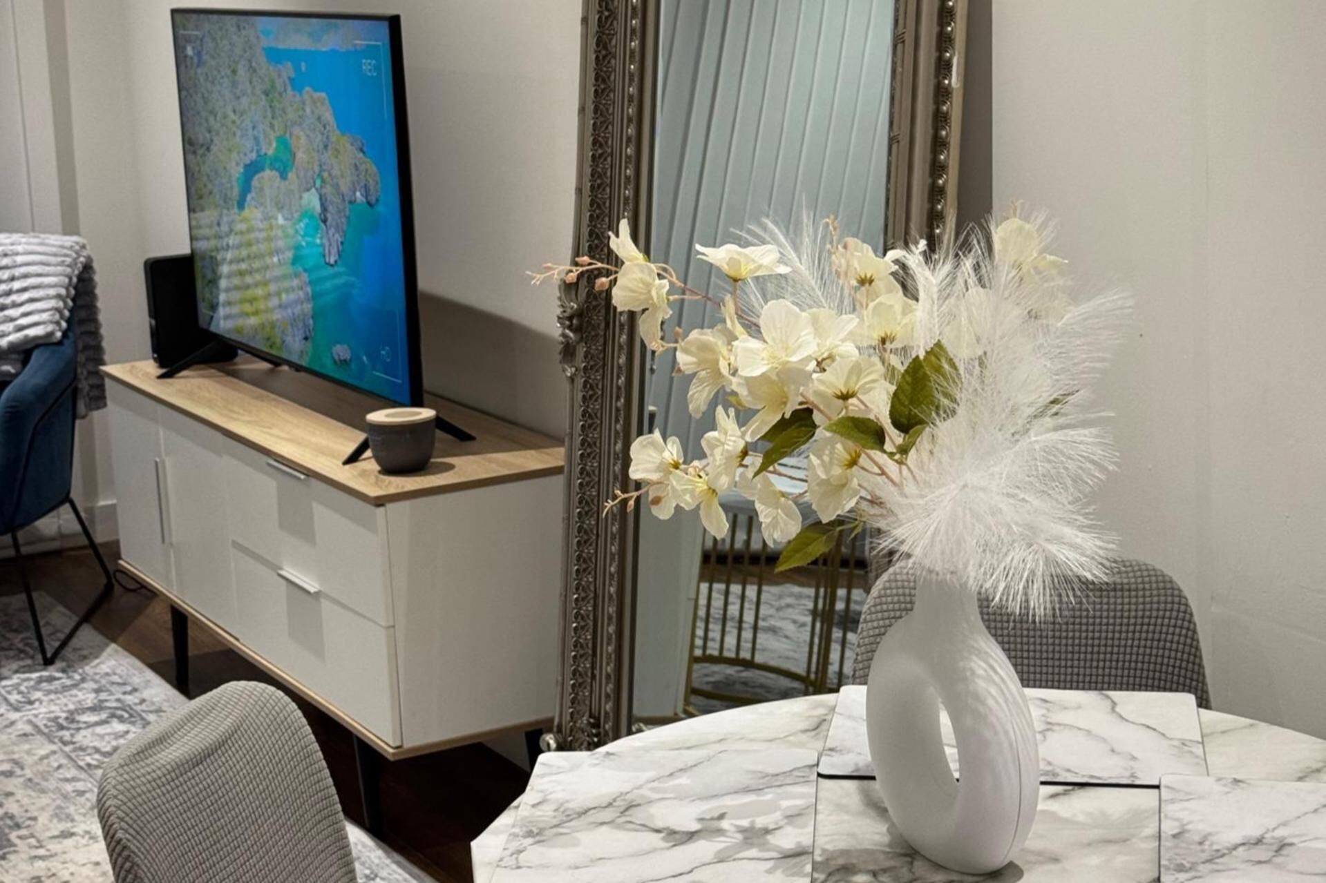 Ein modernes Wohnzimmer mit Fernseher auf einem Holzboden, einer wei ßen Vase mit weißen Blumen und einem Marmortisch.