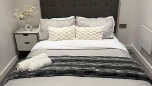 Un lit bien fait, recouvert d’une couverture à rayures grises et blanches, avec des oreillers blancs et un vase blanc rempli de fleurs posé sur une table de chevet.