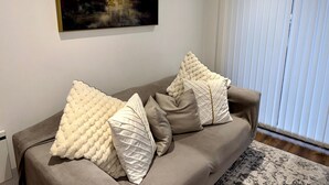 Un salon avec un canapé beige, des coussins décoratifs blancs à texture, un grand tableau abstrait et une fenêtre avec des stores.