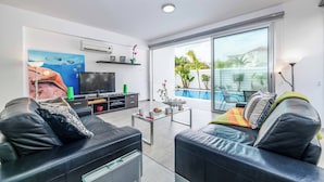 Een moderne woonkamer met twee zwarte leren banken, een glazen salontafel, een flatscreen-televisie en een groot raam met uitzicht op een zwembad en groen.