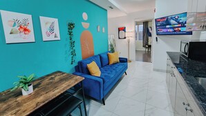 A modern living room with a blue sofa, a wooden dining table, and a kitchen area with a TV.