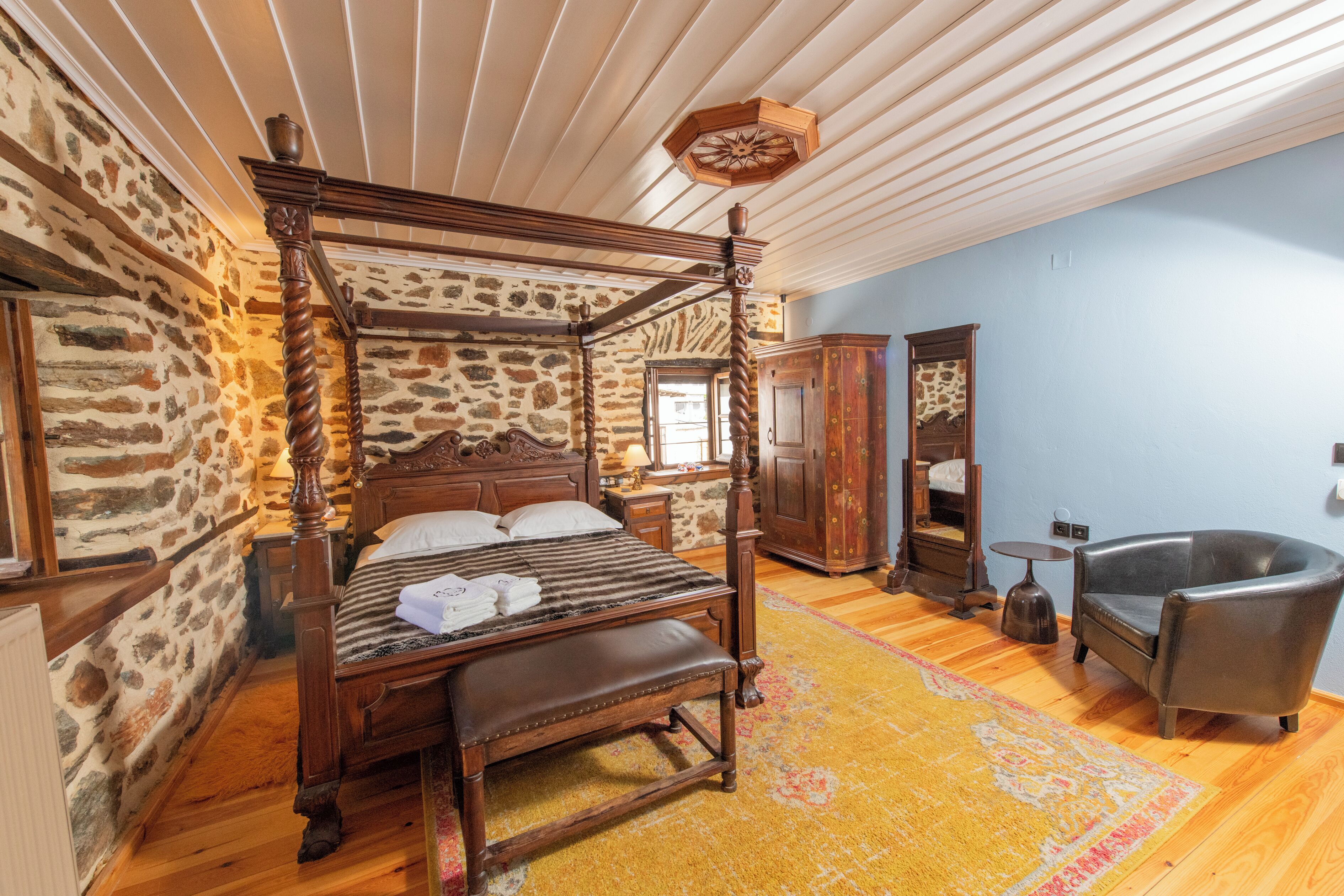 A four-poster bed in a room with stone walls, a wooden ceiling, and a large armchair.