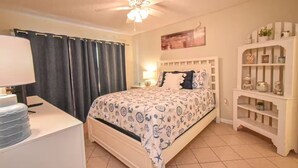Un dormitorio con cama, mesita de noche, lámpara, ventilador de techo, ventana con cortinas y estante con objetos decorativos.