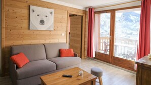 Een gezellige kamer met een grijze bank, rode kussens, een houten tafel en een groot raam met uitzicht op bergen.