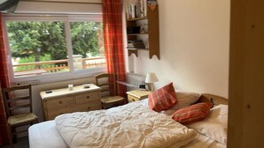 Een slaapkamer met een bed, een bureau en een stoel. Er is een raam met uitzicht op bomen en een hek.