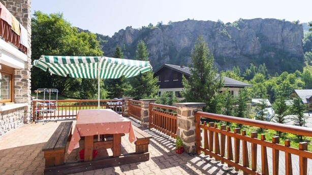Een balkon met een tafel en stoelen, een gestreepte parasol en uitzicht op een berg en bomen.