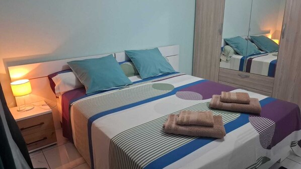 A neatly made bed with striped bedding and two folded towels, a bedside lamp, and a mirror reflecting the bed.
