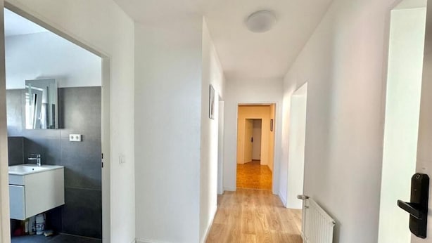 A hallway with wooden flooring, white walls, and a door leading to a bathroom with a white sink and dark tiles. - Family Apartments Near City Centre (Frankfurt)