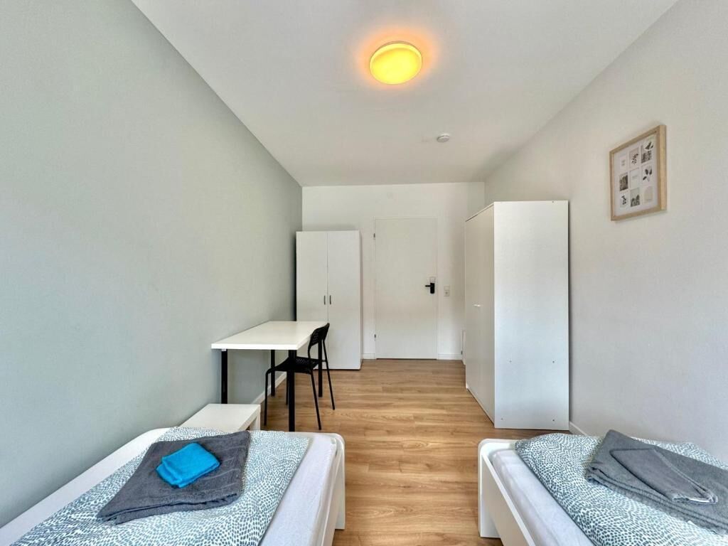 Une petite chambre moderne, équipée de deux lits, d’un bureau et d’une chaise. Une porte donne accès à une autre pièce, et un tableau encadré orne le mur.