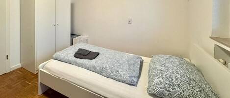 Een kleine kamer met een enkel bed, houten plafond, een kast en een klein nachtkastje.